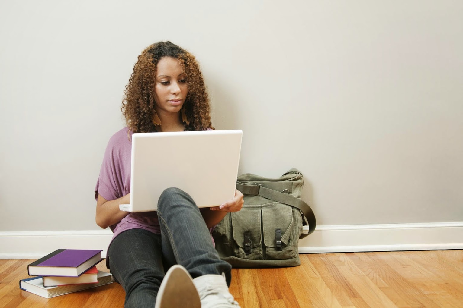 girl sitting on the wooden floor while leaning against a grey wall and looking at her laptop with green tote next to her