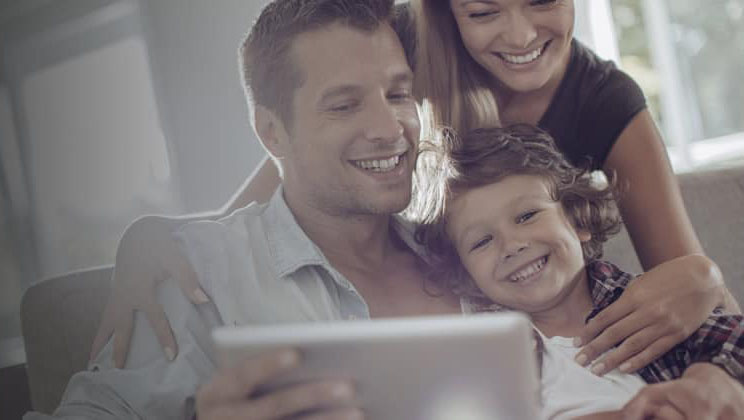 Man, woman, and boy sitting together on a chair looking at tablet in man's hand