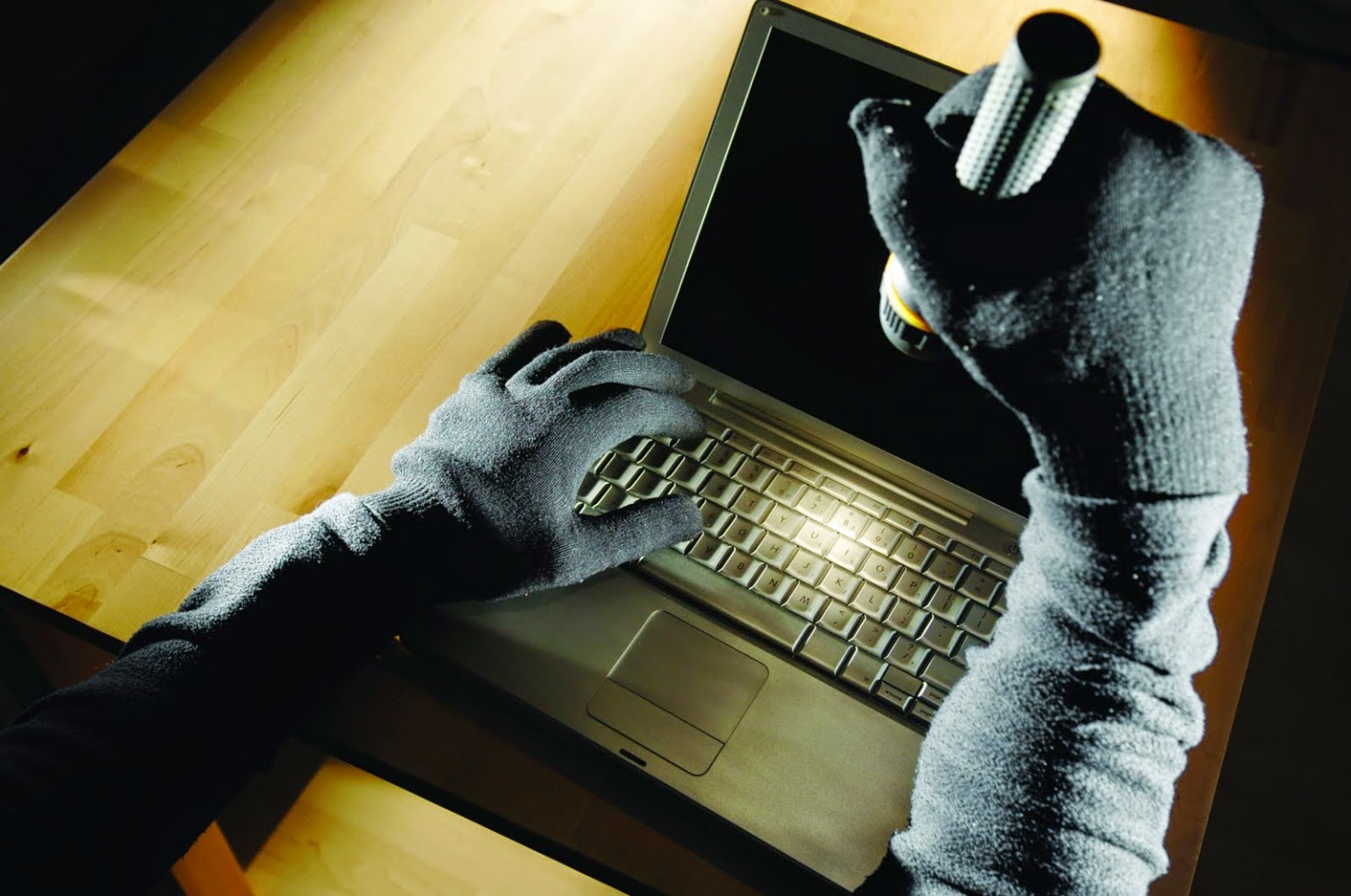 person wearing all black sweatshirt and gloves while holding a flashlight over a laptop