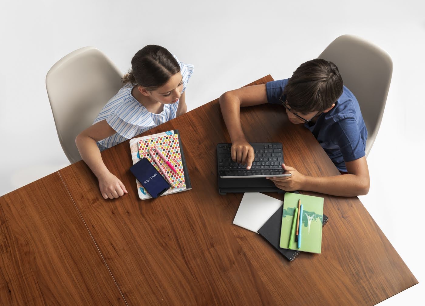 girl and boy sitting at a table looking at a tablet screen with notebooks on the table